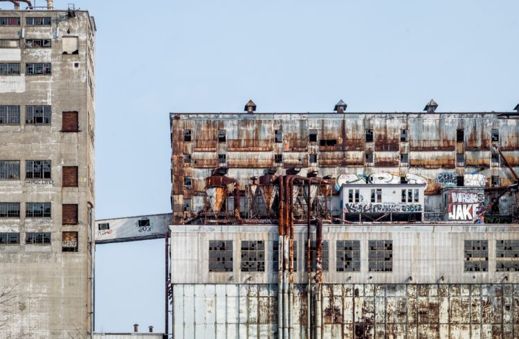 picture of two buildingswith rust all over, graffiti and broken windows against a blue sky