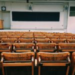 view of an empty university lecture hall with old wooden chairs looking in from the back toward the front with a big chalkboard