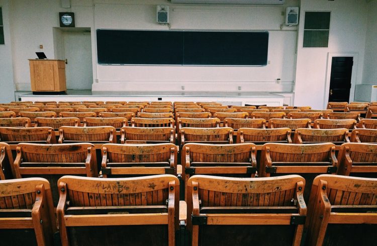 view of an empty university lecture hall with old wooden chairs looking in from the back toward the front with a big chalkboard