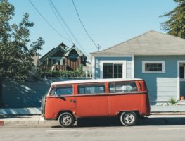 Old, red, vw van outside of a blue house in a neighborhood