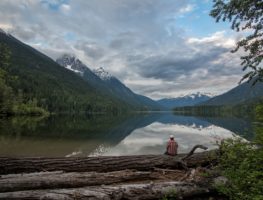 man sitting on a fallen tree branch looking out at a river and mountains