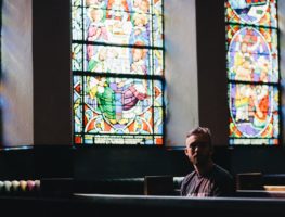 20-something young white man sitting in a pew in a church with stained glass windwos