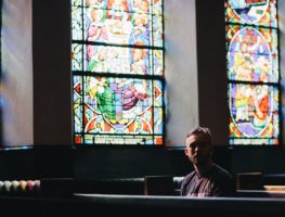 20-something young white man sitting in a pew in a church with stained glass windwos