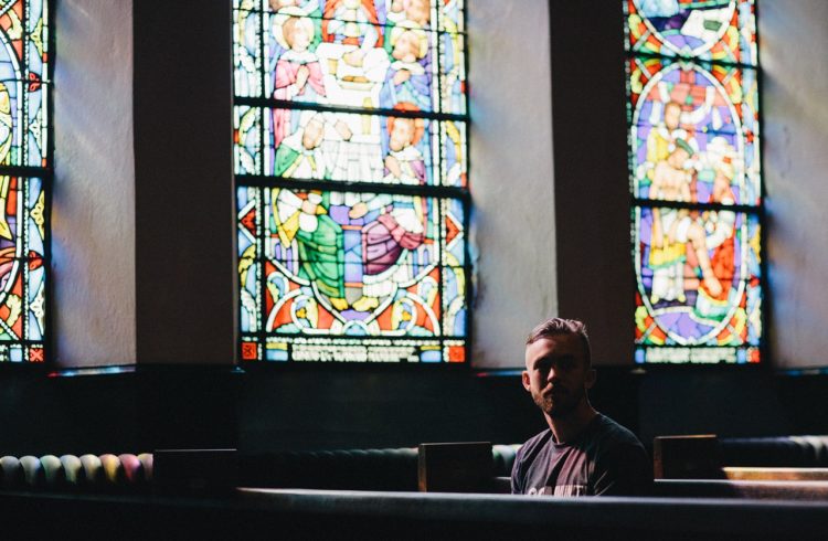 20-something young white man sitting in a pew in a church with stained glass windwos