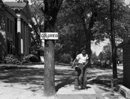black girl stands at a water fountain with the sign 'colored' posted on the tree next to it