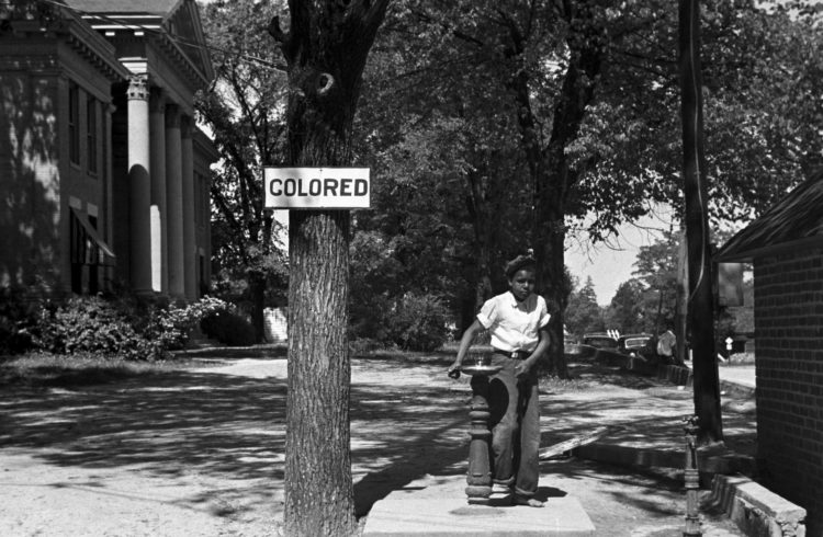 black girl stands at a water fountain with the sign 'colored' posted on the tree next to it
