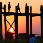 teek bridge in the foreground of a pink and orange sunset with people on the bridge as silouhettes