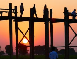 teek bridge in the foreground of a pink and orange sunset with people on the bridge as silouhettes