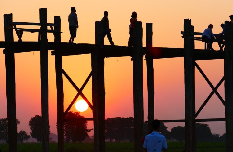 teek bridge in the foreground of a pink and orange sunset with people on the bridge as silouhettes