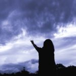 silouhette of a woman raising her hand to a dark blue cloudy sky