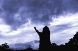 silouhette of a woman raising her hand to a dark blue cloudy sky