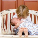 blonde toddler holds baby in arms on an outdoor, white bench in front of an orange door