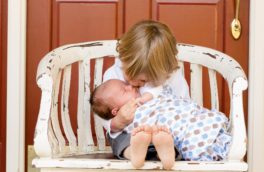 blonde toddler holds baby in arms on an outdoor, white bench in front of an orange door